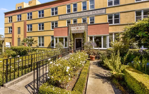 Exterior view of a yellow brick senior living facility with a paved entrance and garden.