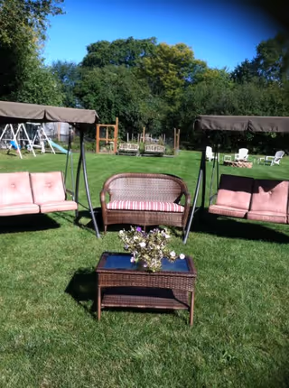 Outdoor seating area on a grassy lawn with two cushioned swings and a wicker loveseat with a striped cushion. In front of the seating is a wicker coffee table with a glass top and a flower arrangement. Trees and garden structures are visible in the background under a clear blue sky.