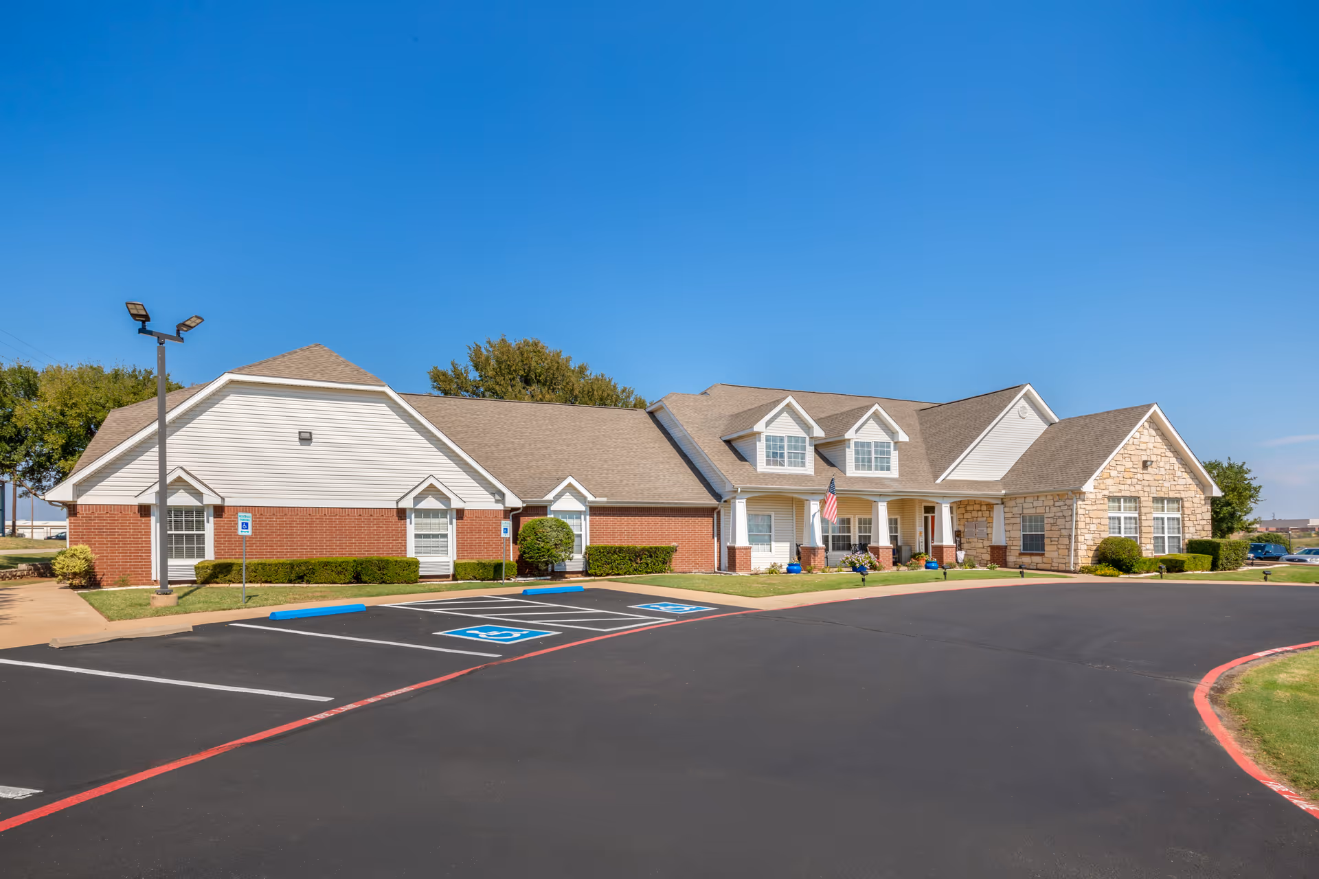 Exterior view of a single-story senior living facility building with a combination of brick, stone, and siding. The building has multiple windows, a covered entrance with white columns, and an American flag displayed near the entrance. There is a paved parking lot in front with marked handicap parking spaces and a clear blue sky overhead.