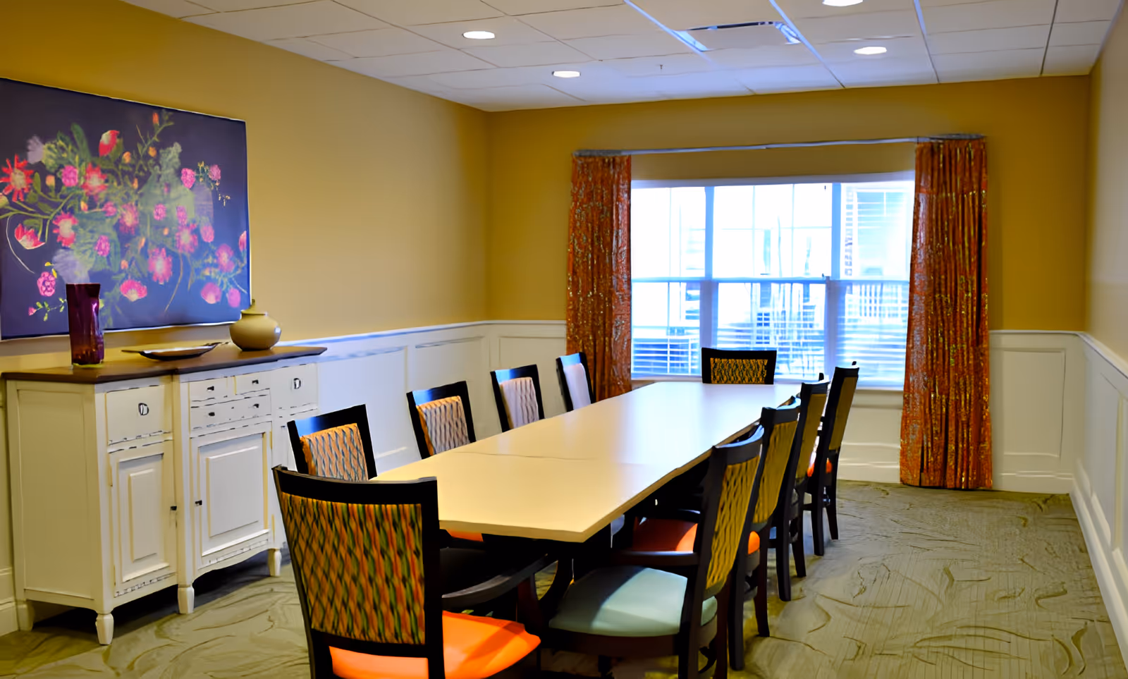 A dining room with a long rectangular table surrounded by ten chairs with colorful cushions. There is a white sideboard against the left wall with decorative items on top and a large floral painting above it. A window with orange patterned curtains is at the far end of the room, letting in natural light. The walls are painted yellow with white wainscoting, and the ceiling has recessed lighting.