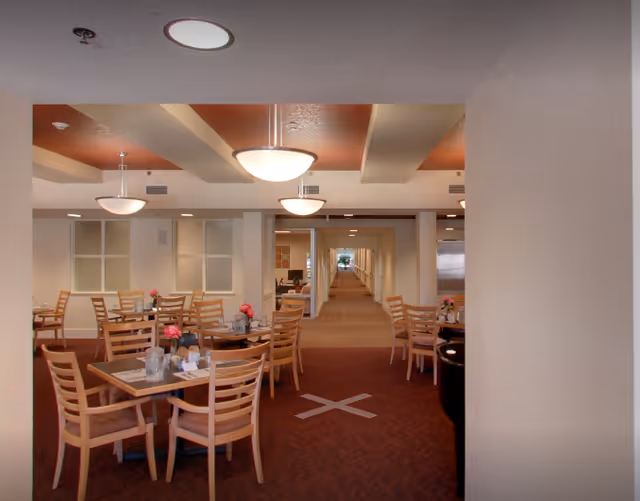 Dining area with wooden tables and chairs, pendant lights overhead, and a long corridor visible in the background.