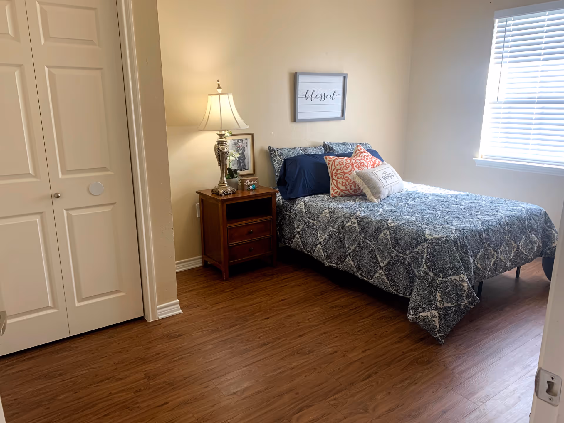 A bedroom with a bed covered in a blue and white patterned quilt, decorated with multiple pillows including a red patterned one and a white one with text. There is a wooden nightstand beside the bed with a lamp, a framed photo, and a small plant. A framed sign with the word 'blessed' hangs on the wall above the bed. The room has wooden flooring and a window with blinds letting in natural light. A white closet door is visible on the left side.