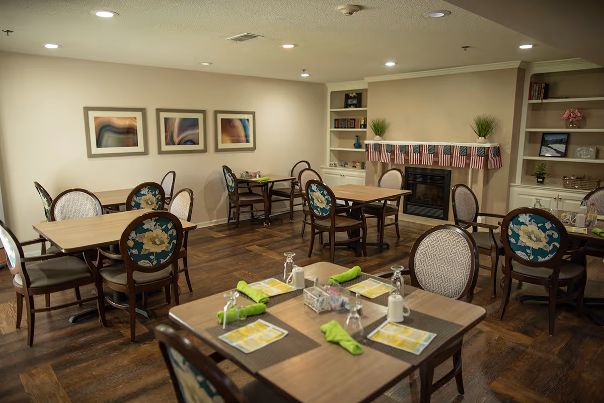 A dining room in an assisted living facility with several wooden tables and chairs arranged neatly. Each table is set with placemats, menus, glasses, and green cloth napkins. The room has wooden flooring, a fireplace decorated with small American flags, built-in shelves with books and plants, and three abstract paintings on the wall.