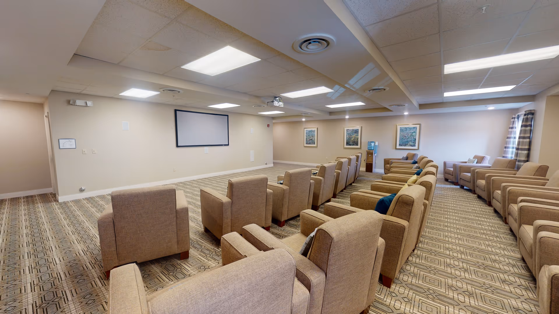 A spacious room with multiple rows of beige armchairs arranged facing a large blank screen mounted on the wall, with patterned carpet flooring, ceiling lights, and framed artwork on the walls.