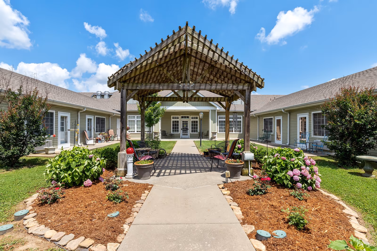 Outdoor courtyard area at Brookfield Assisted Living featuring a wooden pergola with seating underneath, surrounded by landscaped flower beds with blooming hydrangeas and shrubs, and a pathway leading to the building entrance under a bright blue sky with scattered clouds.