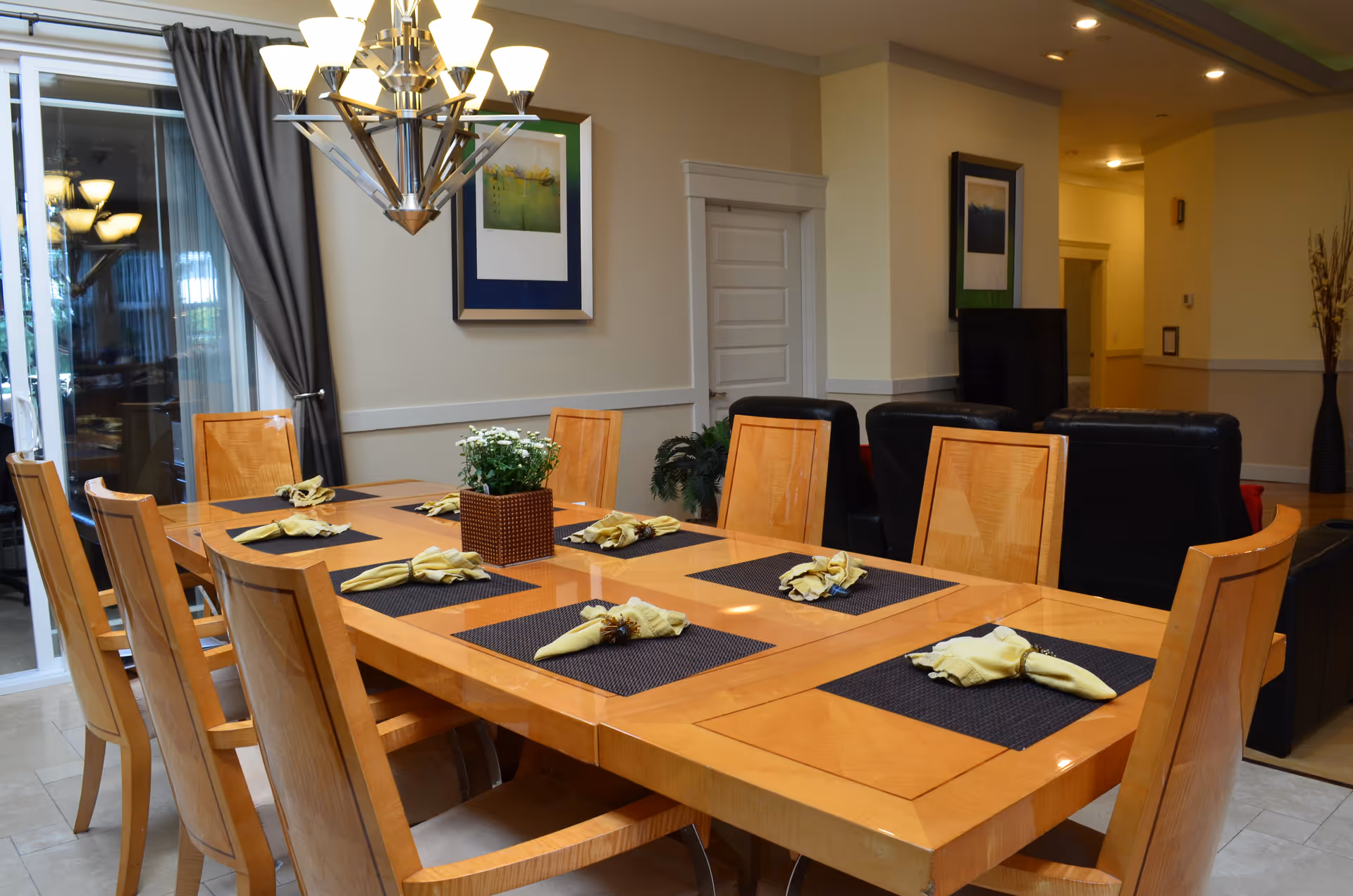 A dining room with a large wooden table set with eight place settings, each with a black placemat and a folded yellow napkin. A modern chandelier hangs above the table. In the background, there are black leather chairs and framed artwork on the walls. A sliding glass door with gray curtains is visible on the left side.
