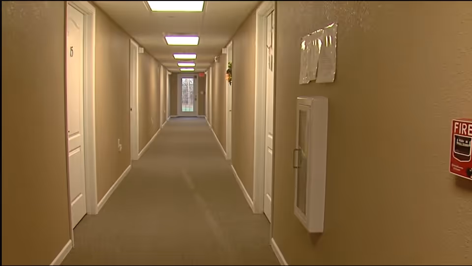 A long carpeted interior hallway in a residential care facility with closed doors on both sides and ceiling lights leading to a door at the far end.