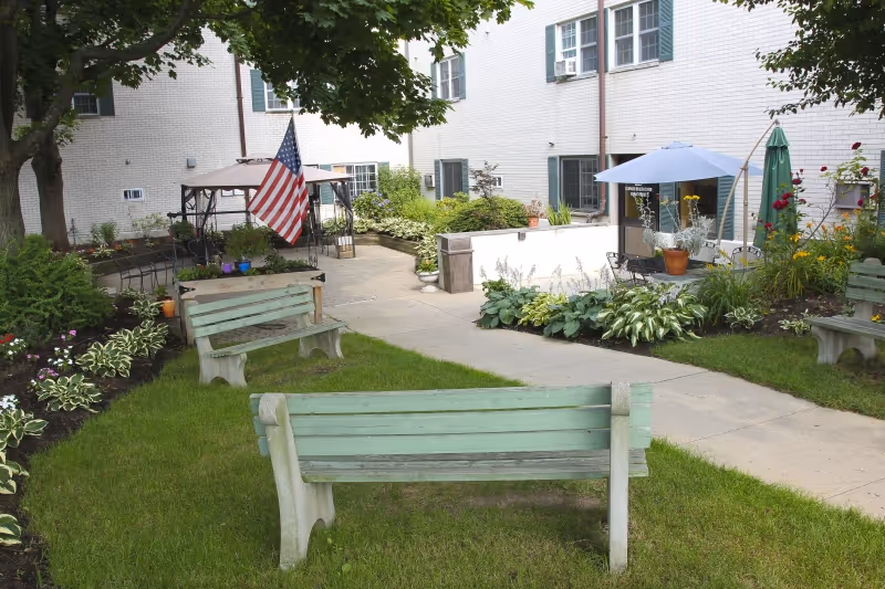 Outdoor garden area at Elmwood Nursing & Rehabilitation Center with green benches, a paved walkway, various plants and flowers, an American flag, and a white building with windows in the background.