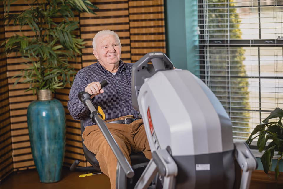 An elderly man with white hair wearing a checkered shirt and brown pants is sitting on a rowing exercise machine indoors. Behind him is a large blue vase with a green leafy plant and a window with blinds letting in natural light.