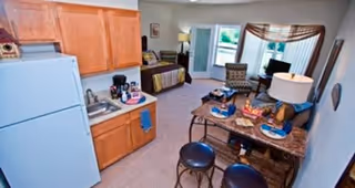 Interior view of a retirement living unit showing a small kitchen area with wooden cabinets, a refrigerator, and a sink. Adjacent to the kitchen is a dining table set for two with plates and glasses. Beyond the dining area is a living space with a chair, a bed, and a window with curtains letting in natural light.