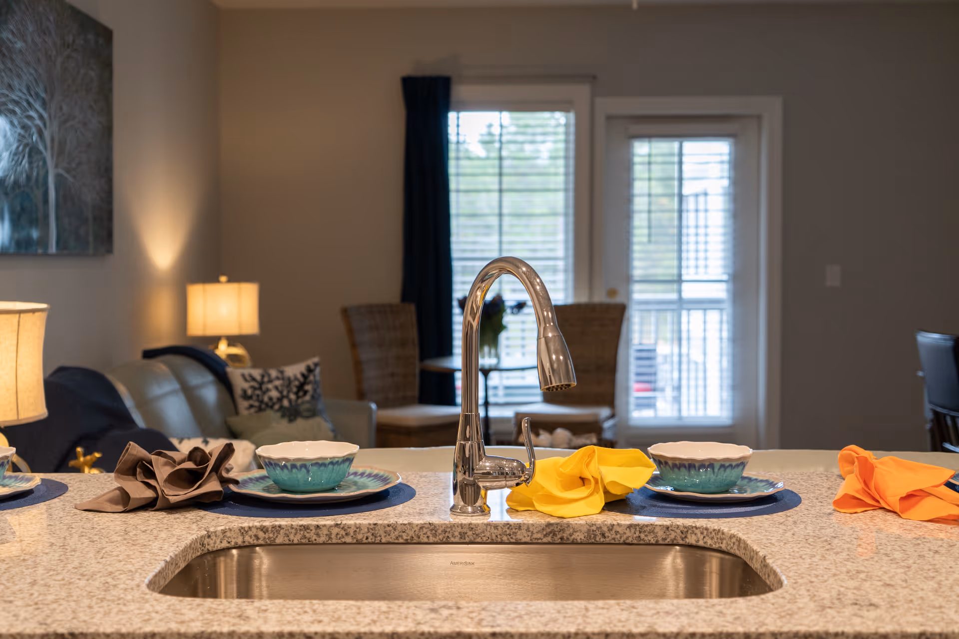 View of a kitchen sink with a modern chrome faucet on a granite countertop. The countertop has place settings with bowls, plates, and colorful napkins. In the background, there is a living area with a sofa, cushions, a lamp, and a window with blinds and a door leading outside.
