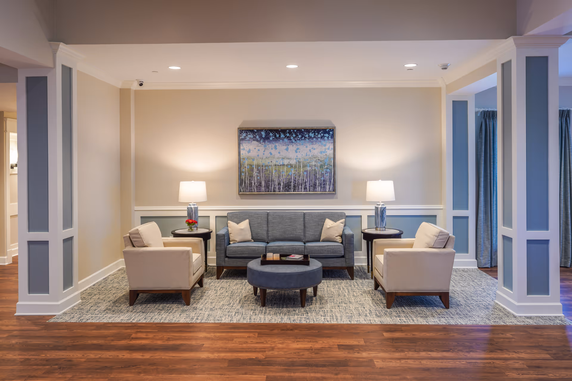 A cozy living room area with a gray sofa, two beige armchairs, a round ottoman coffee table, and two side tables with lamps. A colorful abstract painting hangs on the wall behind the sofa. The room has wooden flooring and a large area rug, with soft lighting creating a warm atmosphere.