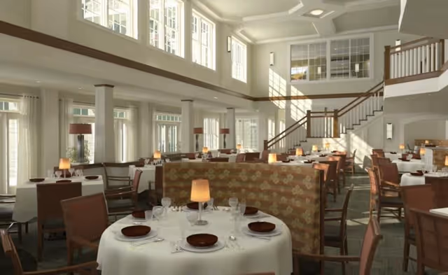A spacious and elegant dining room with multiple round and rectangular tables set with white tablecloths, brown plates, glassware, and small lamps. The room features large windows allowing natural light, high ceilings, and a staircase leading to an upper level.