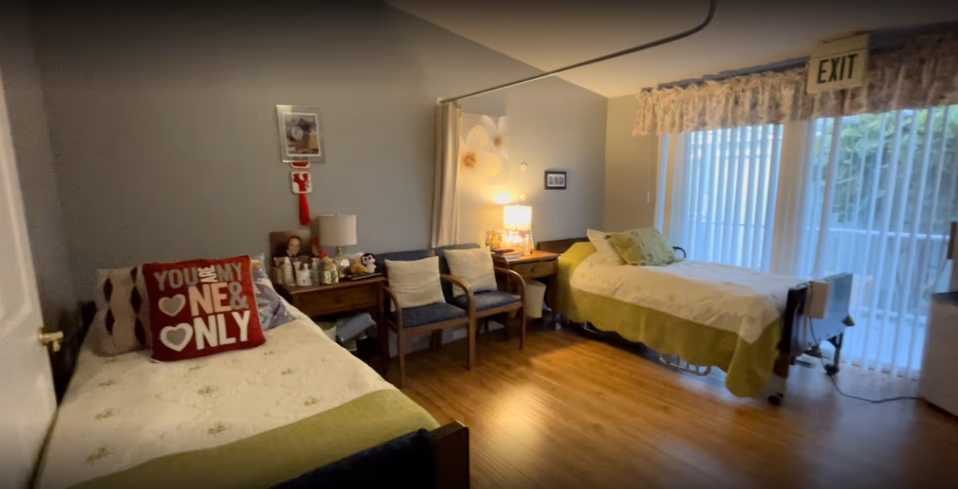 Shared senior bedroom with two twin beds, chairs and a nightstand beside a sliding glass door with vertical blinds and an EXIT sign.