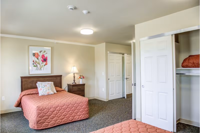 A bedroom in a senior living facility with two beds covered in matching coral quilted bedspreads. There is a wooden nightstand between the beds with a lamp and a small vase of flowers. A framed floral artwork hangs on the wall above one bed. The room has beige walls, carpeted floor, and a closet with sliding white doors, one of which is open showing a folded coral blanket inside.