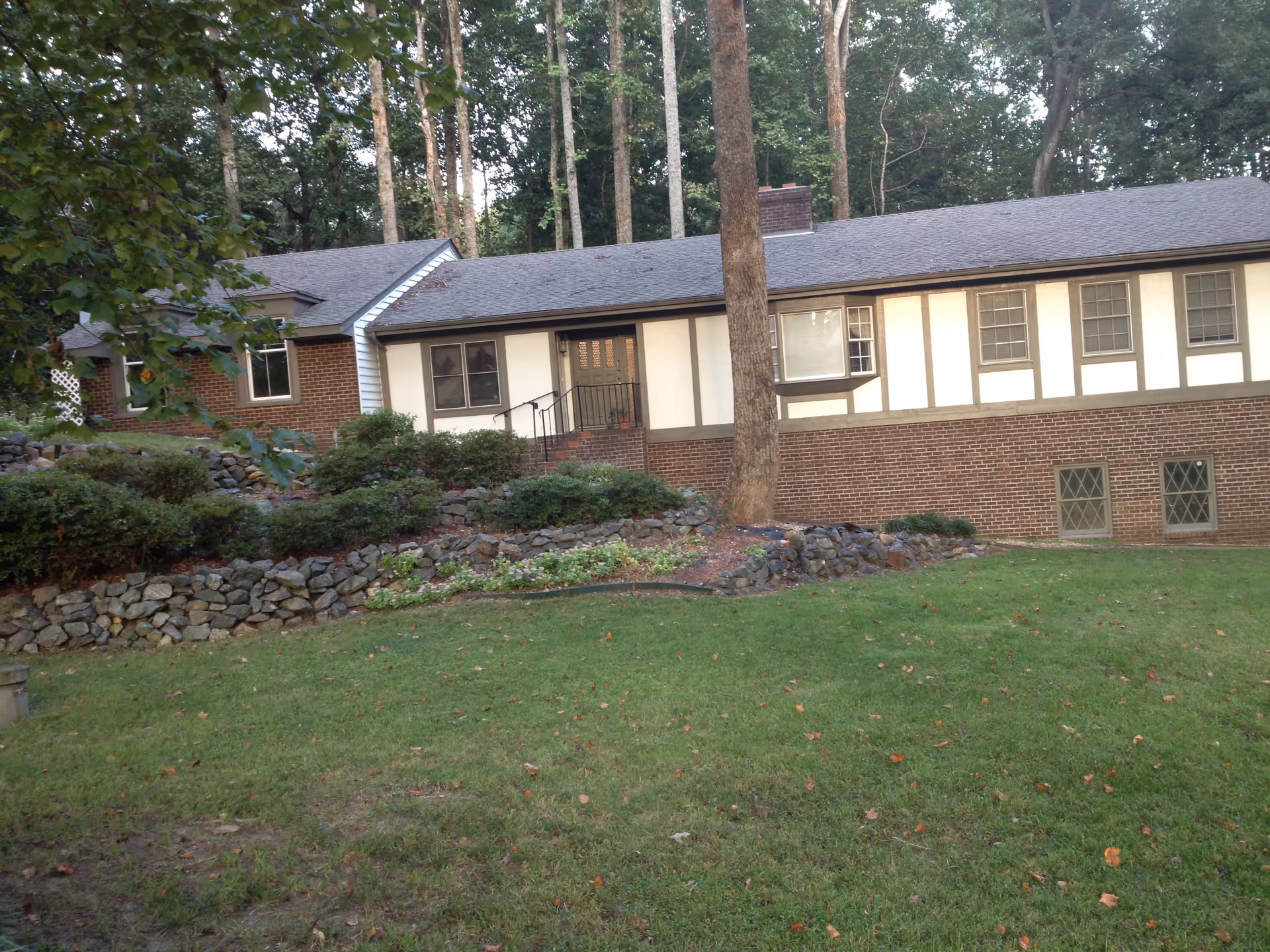 Exterior view of a single-story assisted living facility building with a brick lower half and white upper half with brown trim. The building is surrounded by a grassy lawn, stone retaining walls, bushes, and tall trees in the background.
