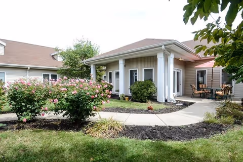 Exterior view of a single-story building with beige siding and a brown roof, featuring a covered porch supported by white columns. There is a paved walkway curving through a landscaped garden with green grass, bushes, and pink flowering plants. Outdoor seating with a table and chairs is visible under a red awning on the right side.