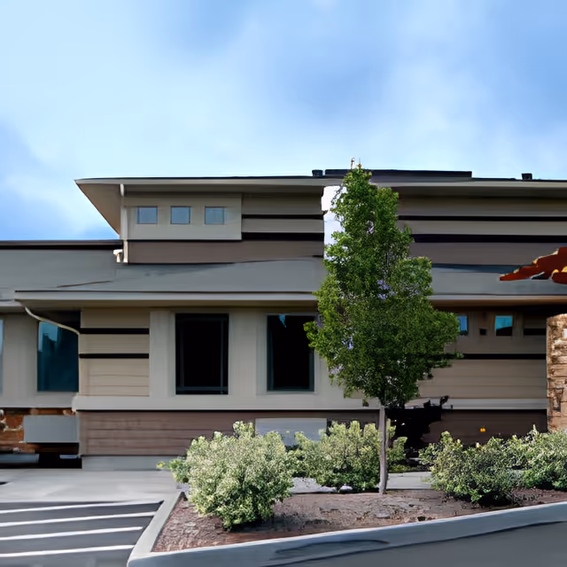 Front exterior of a modern assisted living building with windows, a small landscaped bed, and a young tree.