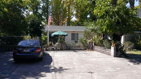 Front exterior of Vintage Chalet senior living, driveway, parked car, and shaded patio with umbrella.