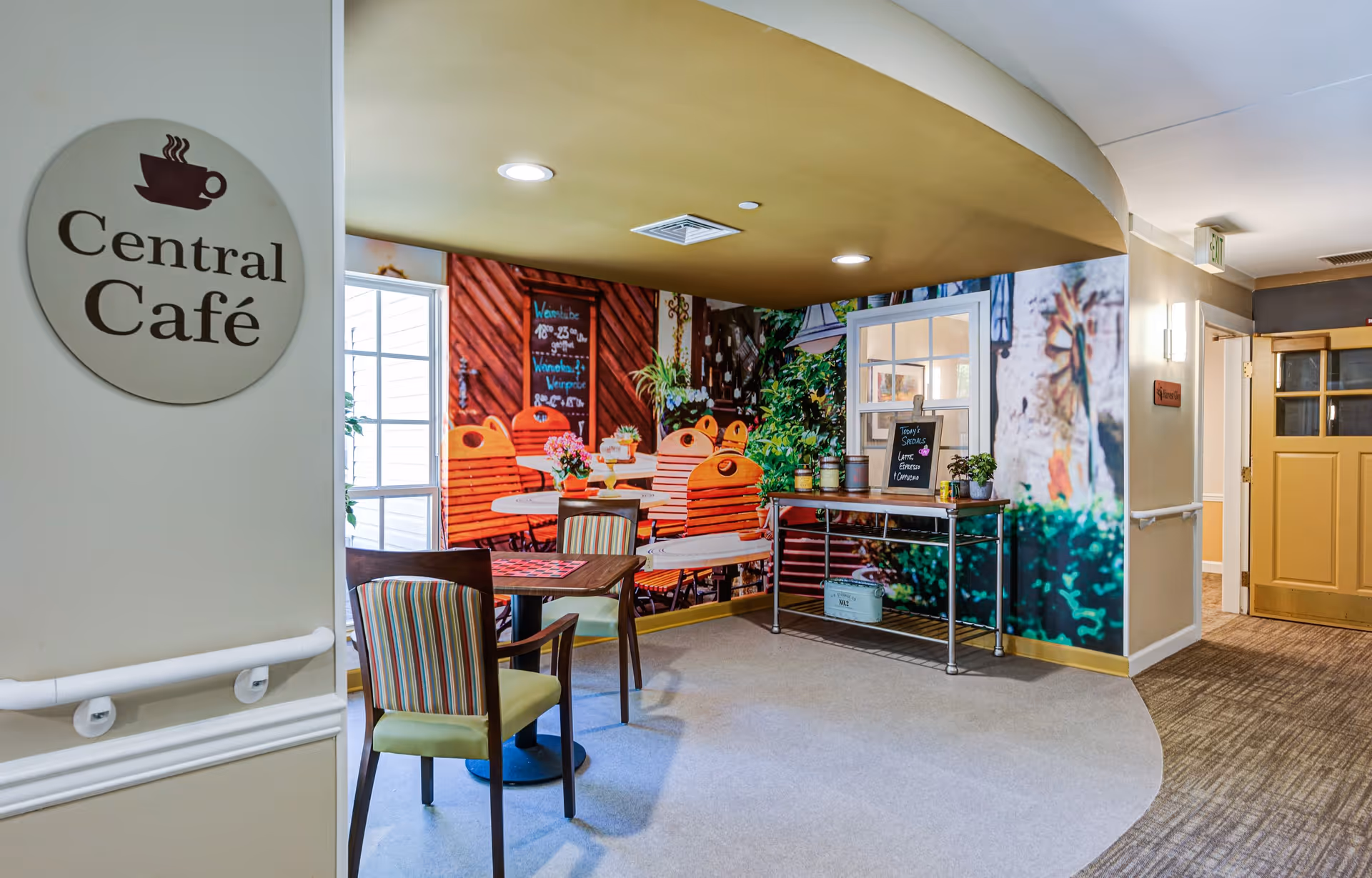 Interior view of a cozy café area named Central Café with a round table and two chairs. The wall features a large mural depicting an outdoor café scene with wooden chairs, plants, and a menu board. There is a small table with potted plants and a chalkboard sign listing daily specials. The space is well-lit with recessed ceiling lights and has a warm, inviting atmosphere.