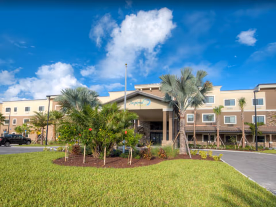Front exterior of a three-story senior living building with a covered entrance, palm trees, and landscaped lawn under a blue sky.
