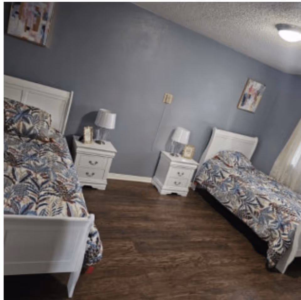A bedroom with two single beds featuring white wooden frames and colorful patterned bedding. Between the beds are two white nightstands, each with a lamp and a small framed picture. The walls are painted gray, and there are two abstract paintings hanging on the walls. The floor is wooden, and a ceiling light is visible.