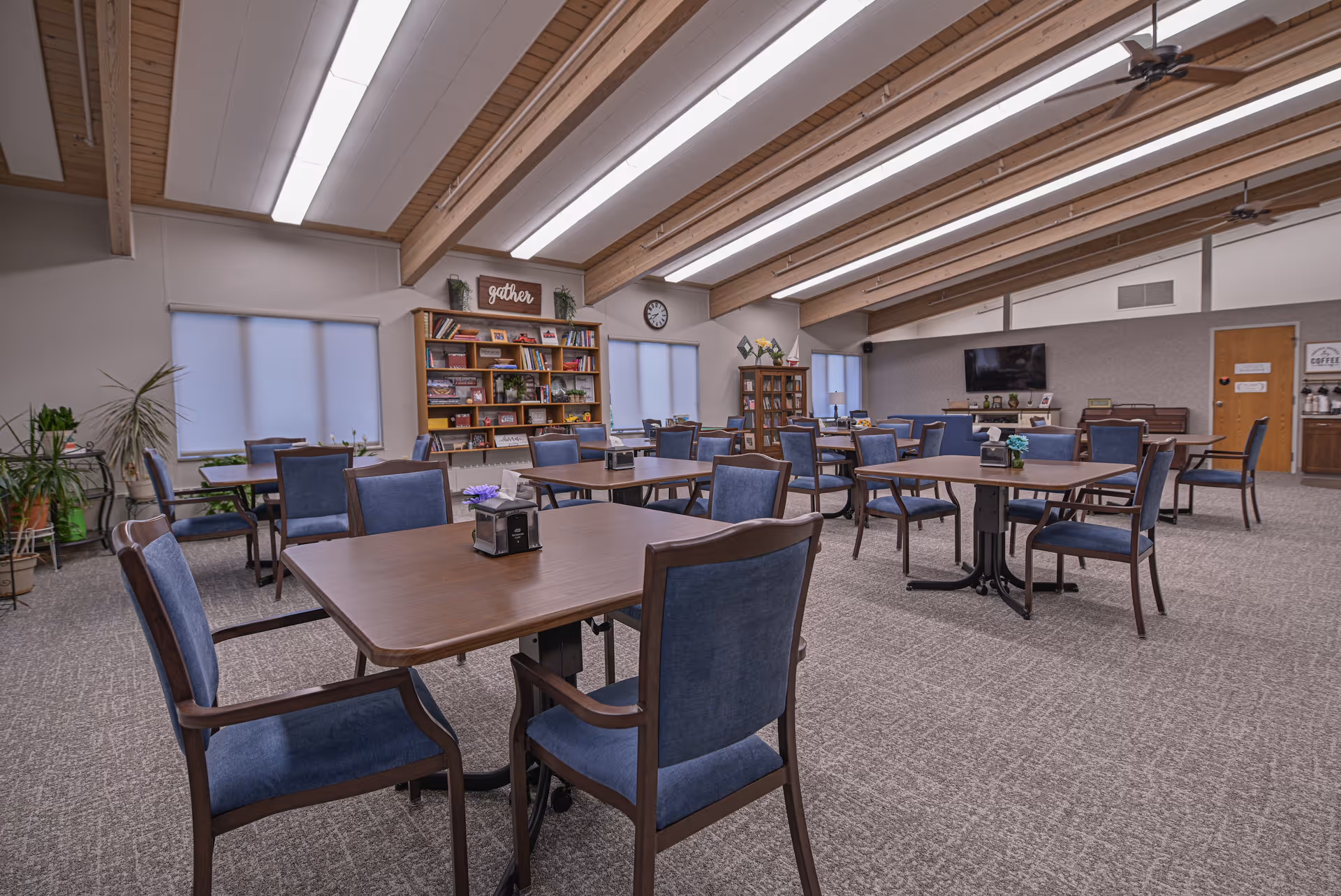 A spacious dining room with multiple wooden tables and blue cushioned chairs arranged neatly. The room has a high ceiling with exposed wooden beams and fluorescent lighting. There are large windows with blinds, a bookshelf filled with books and decorative items, a clock on the wall, and a TV mounted on the far wall. The carpeted floor and overall decor create a warm and inviting atmosphere.