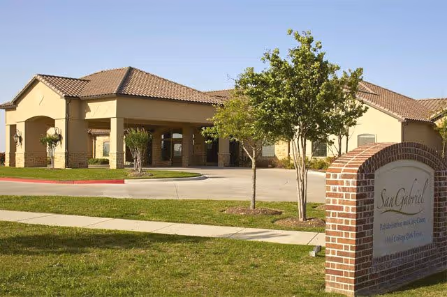 Exterior view of San Gabriel Rehabilitation And Care Center showing a single-story building with a tiled roof, a covered entrance, a driveway, and a brick sign with the facility's name. There are small trees and grass in the foreground under a clear blue sky.