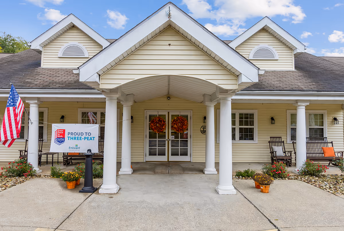 Front entrance of Clen-Moore Place facility with a covered porch supported by white columns, two glass doors decorated with autumn wreaths, an American flag on the left, and outdoor seating with chairs and a bench on the right. There are flower pots and a sign that reads 'Proud to Three-Peat' near the entrance.