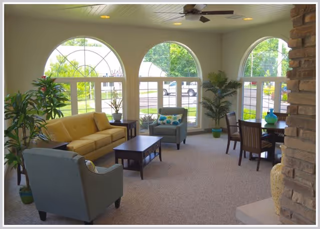 A bright and spacious living room area with large arched windows letting in natural light. The room features a yellow sofa, two blue armchairs, a wooden coffee table, a round dining table with four chairs, several potted plants, and a ceiling fan. The walls are light-colored and there is a stone fireplace partially visible on the right.