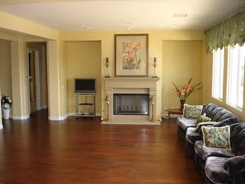 A cozy living room with wooden flooring, a fireplace centered on the far wall with a floral painting above it, two candle holders on the mantel, a small TV on a stand to the left, and a dark upholstered sofa with floral cushions to the right near a window with green valance curtains. A small side table with a flower arrangement is next to the sofa.