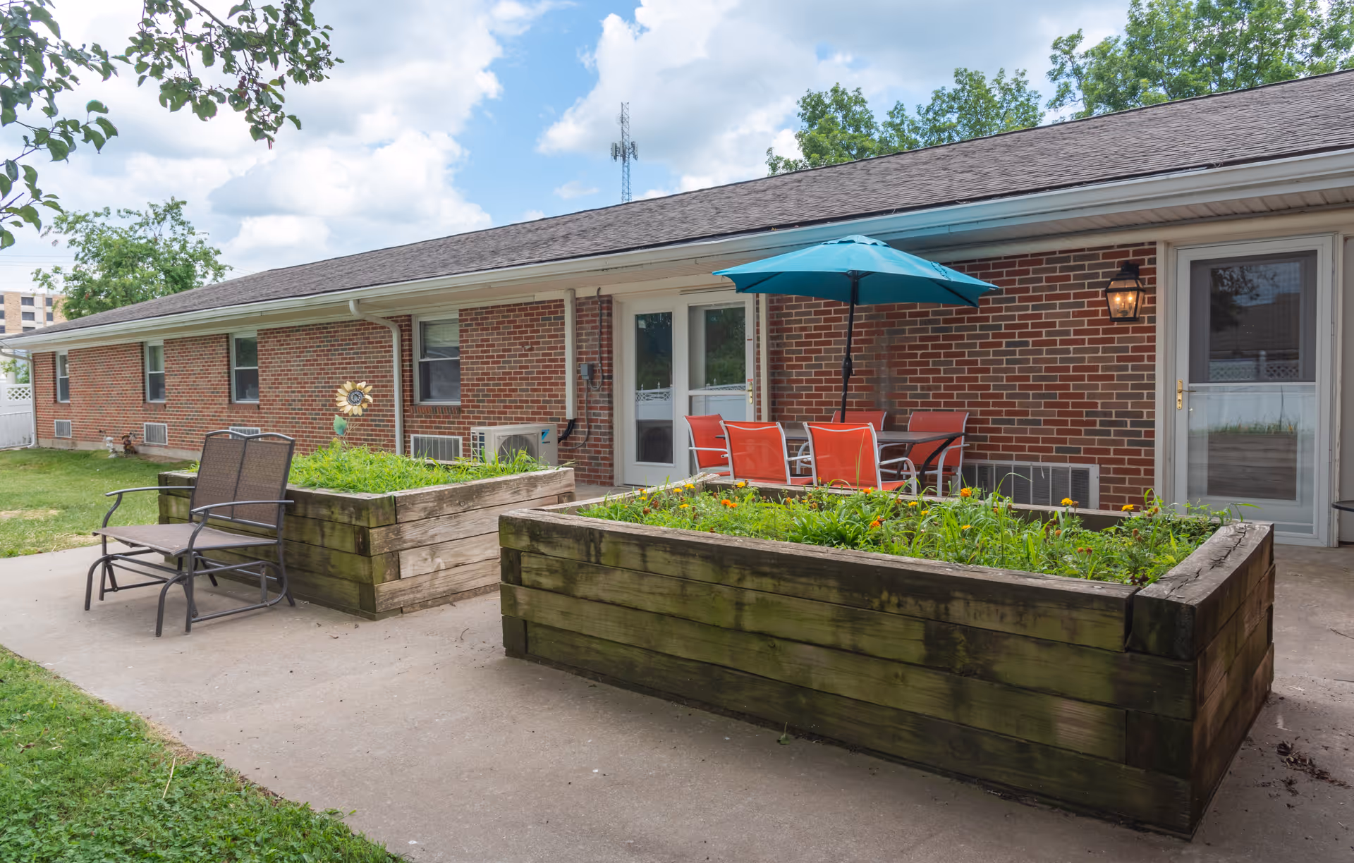 Outdoor patio area at Rolla Presbyterian Manor with raised wooden garden beds filled with plants and flowers, a table with red chairs under a blue umbrella, and two brown chairs on a concrete walkway next to a brick building with white doors and windows.