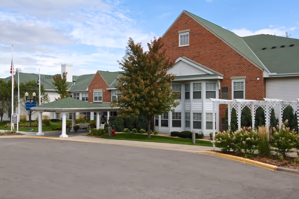 Exterior view of Heathers Manor, a senior living facility with red brick and white siding, green roofs, a covered entrance with white columns, landscaped gardens, and a white trellis on the right side.