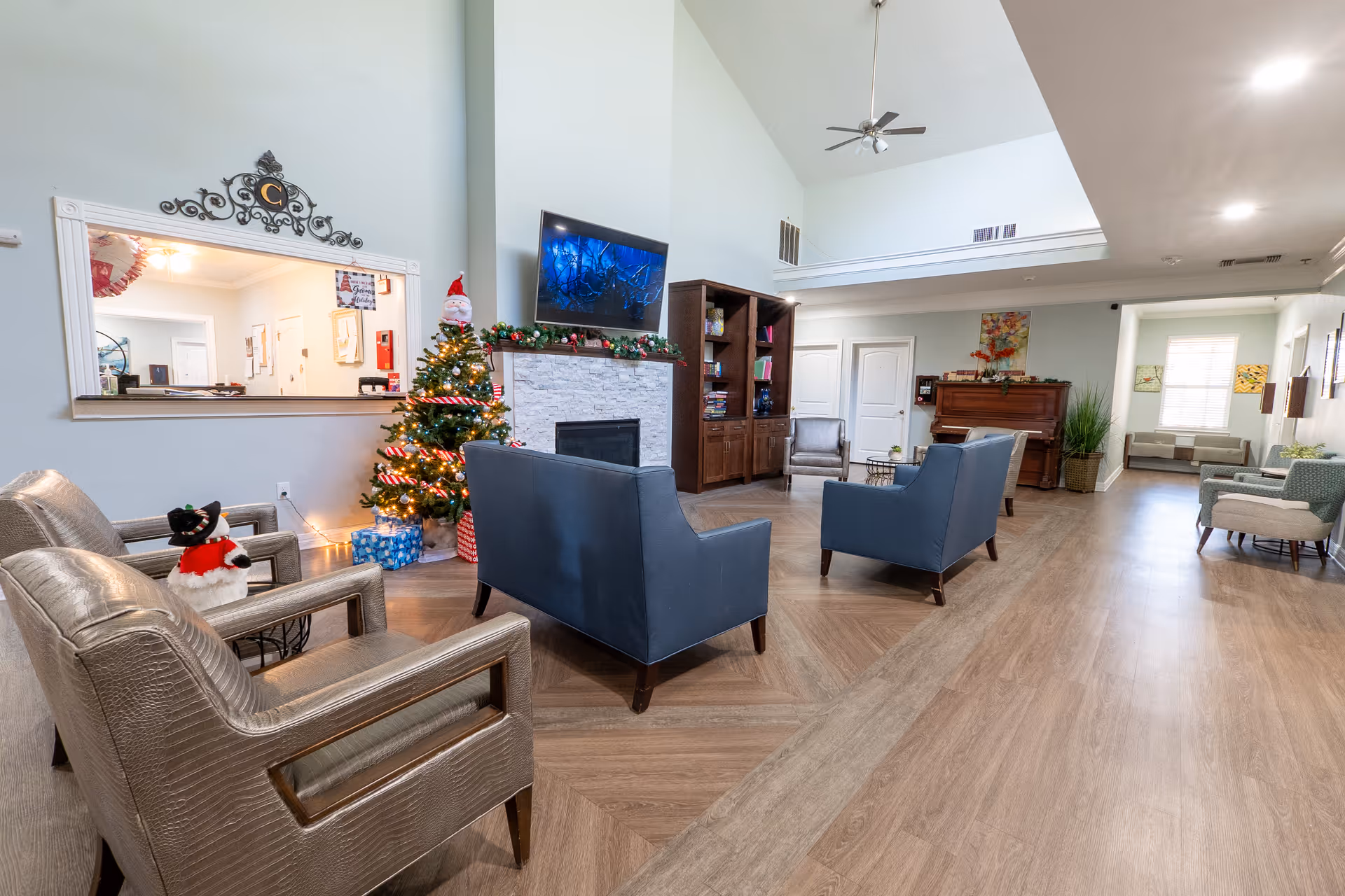 A spacious senior living facility common area decorated for Christmas with a small Christmas tree, presents, and festive garland on the fireplace mantel. The room features comfortable seating including two blue sofas and two beige armchairs, a wall-mounted TV above the fireplace, a wooden bookshelf, and a piano in the background. The space has high ceilings, wood flooring, and large windows allowing natural light.
