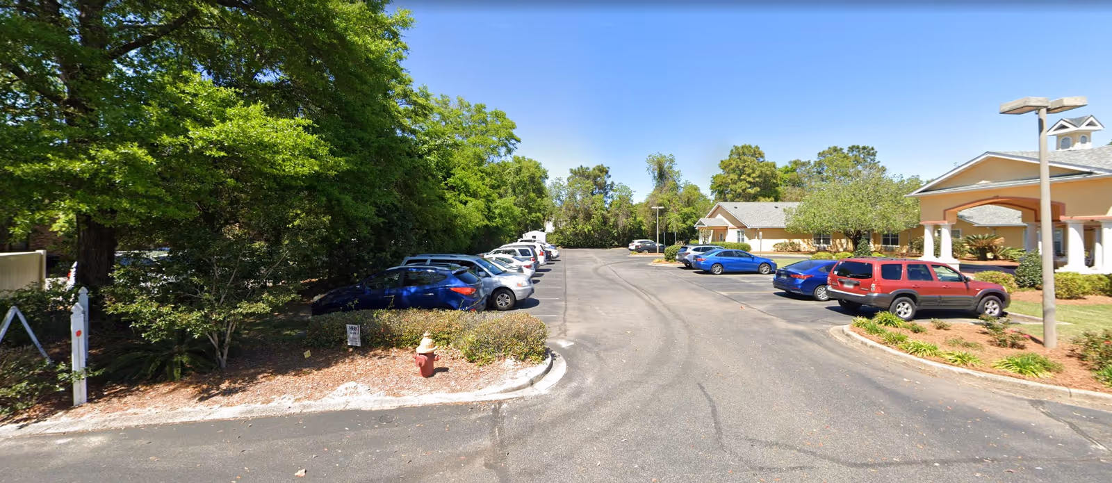 Parking lot and entrance of a senior living facility with parked cars, landscaping, and a single-story building under a clear blue sky.