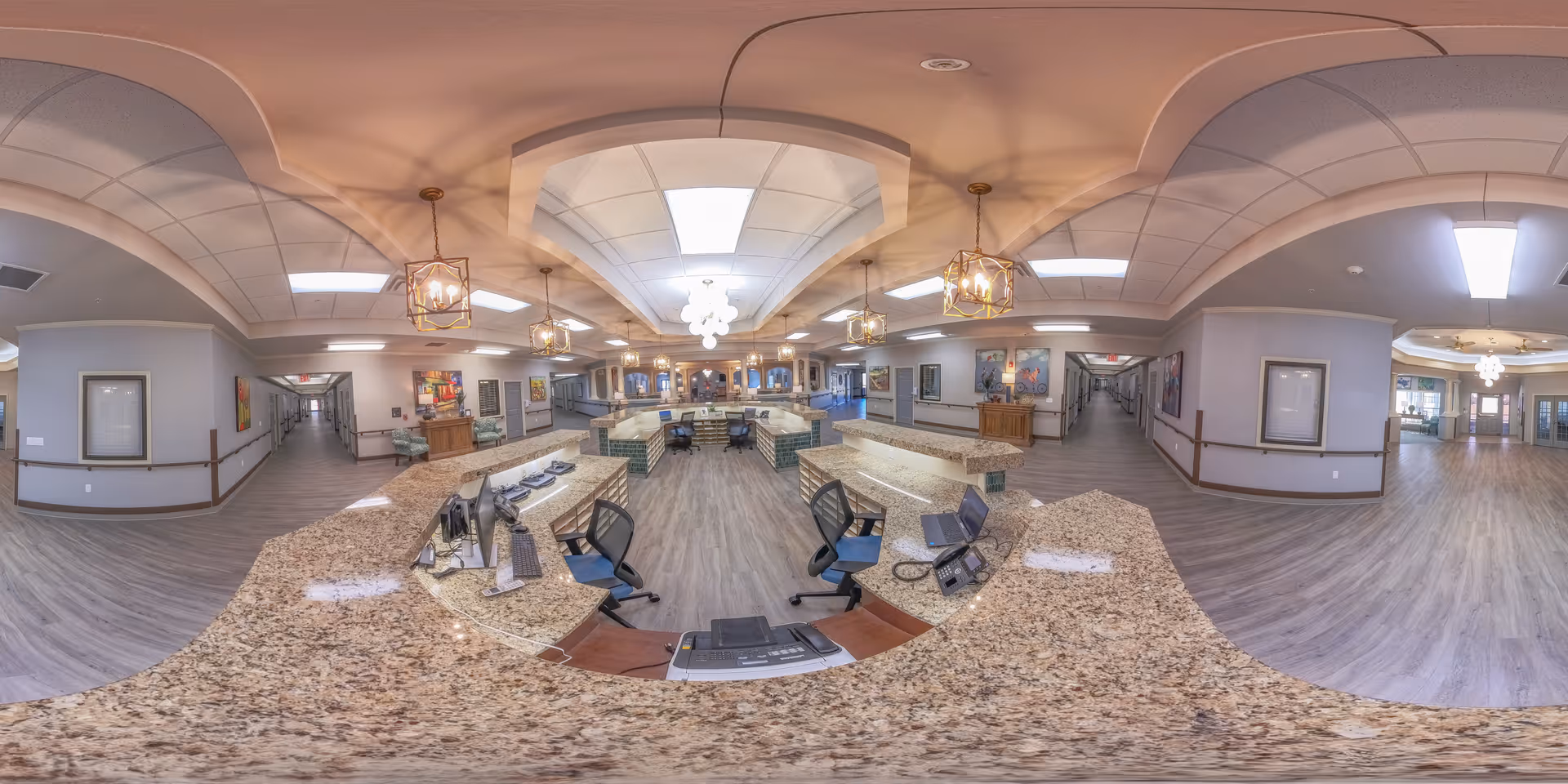 Wide-angle view of a spacious reception area in a senior living facility with a large granite countertop desk, office chairs, pendant lights, and hallways extending on both sides. The floor is wood-style laminate, and the walls are decorated with framed artwork and handrails.