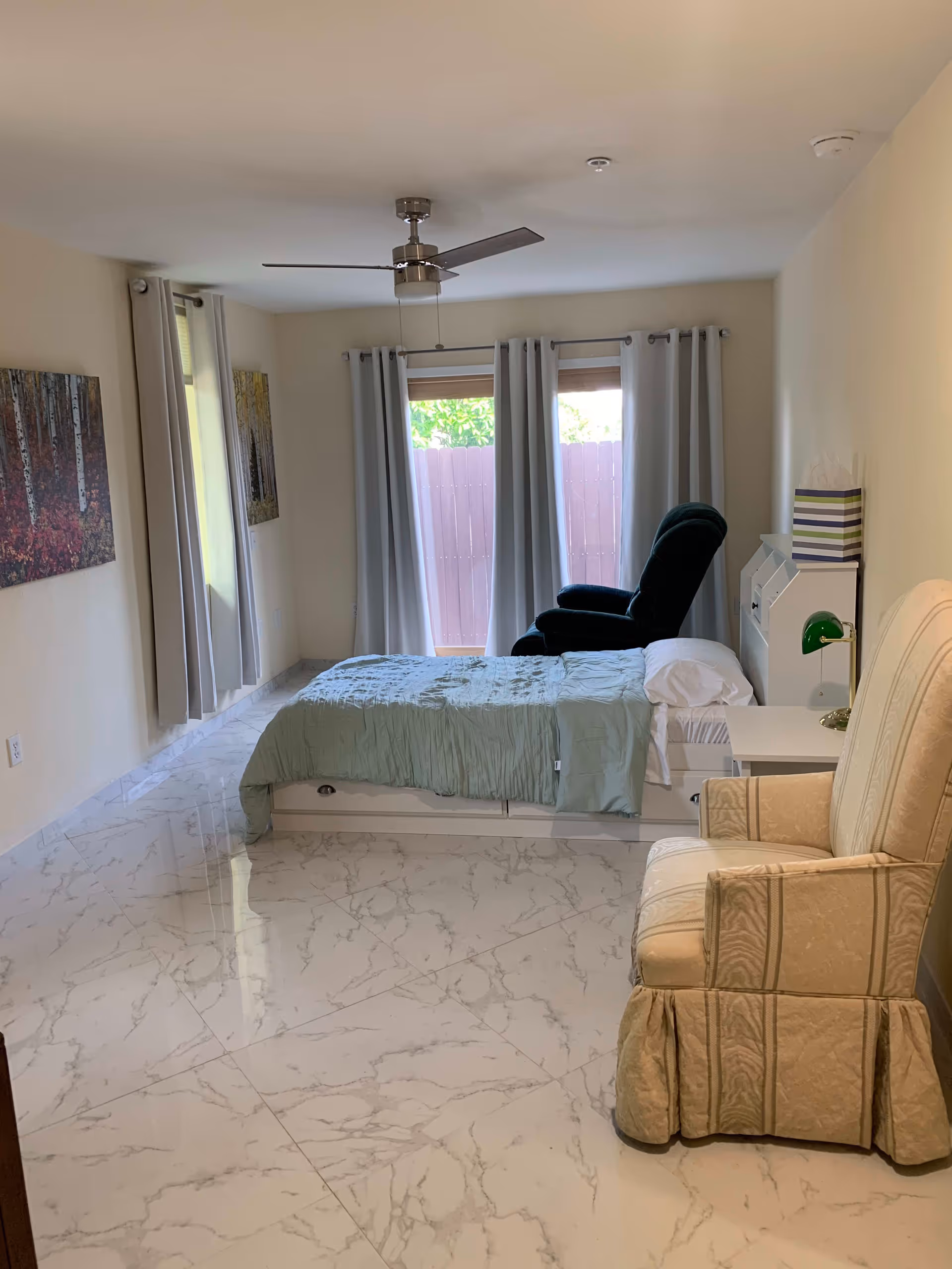 A bright bedroom with a bed covered in a light green comforter, a beige upholstered armchair, a dark green recliner near the window, and two large windows with light gray curtains. The floor is white with gray marble patterns, and there are two paintings on the left wall depicting forest scenes.