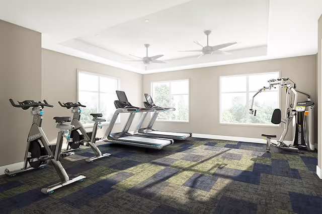 A bright fitness room with two stationary bikes, two treadmills, and a multi-function weight machine. The room has large windows letting in natural light, beige walls, and a patterned carpet floor.