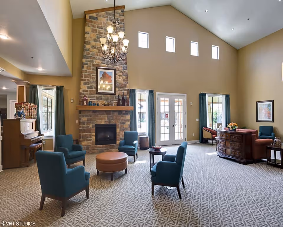 A spacious living room with high vaulted ceilings and a large stone fireplace in the center. Four teal armchairs are arranged around a round brown ottoman and a small table. There is a chandelier hanging from the ceiling. The room has large windows with teal curtains and glass double doors letting in natural light. A piano is visible on the left side near a window, and a wooden dresser with a flower arrangement is on the right side. The carpet has a geometric pattern.