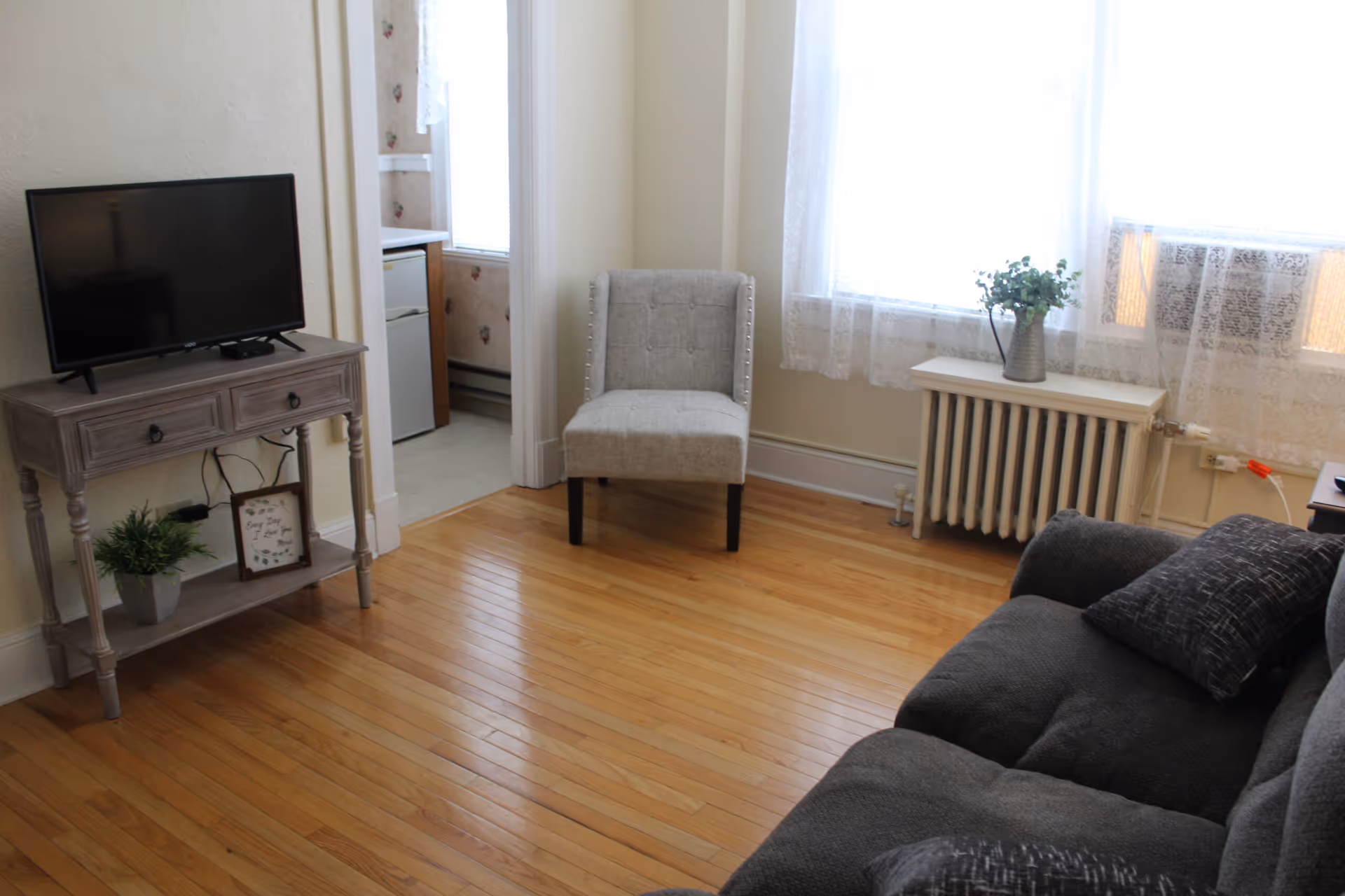 A cozy living room with a gray cushioned armchair, a dark gray sofa with patterned pillows, a small wooden table holding a flat-screen TV, a potted plant, and a framed sign. The room has wooden floors, a radiator under a window with sheer white curtains, and a view into a small kitchen area with a refrigerator.