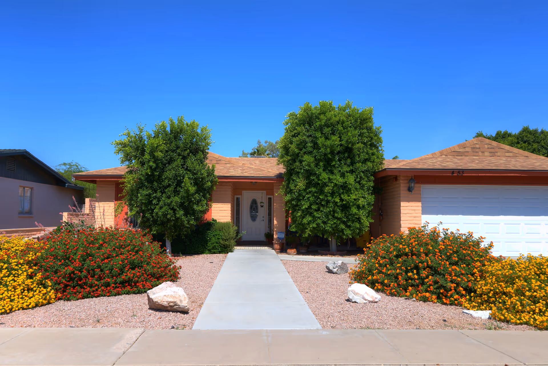 Front exterior of a single-story assisted living facility with a central entrance, two large trees, and colorful landscaping.