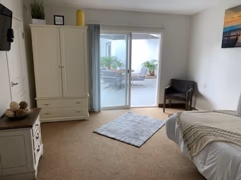 A bright bedroom with a bed on the right side covered with a beige and white blanket, a gray rug on the carpeted floor, a wooden chair in the corner, a white wardrobe and dresser on the left side, and sliding glass doors leading to an outdoor patio with plants and seating.