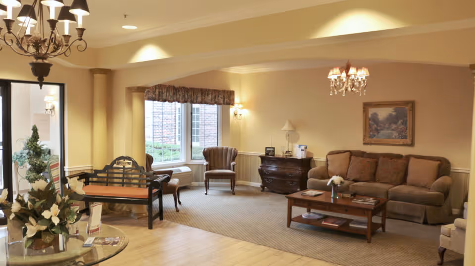 A cozy living room area with a beige sofa, a wooden coffee table with books and a flower vase, an armchair near a window with a valance, a wooden chest with a lamp and framed pictures, and a chandelier hanging from the ceiling. There is also a bench with a cushion and a glass table with a floral arrangement in the foreground.