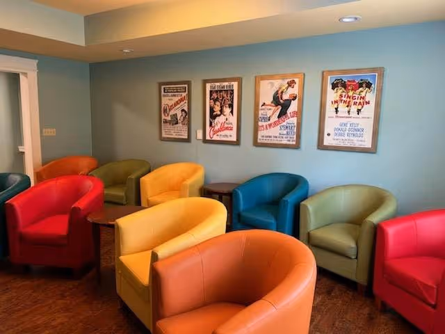 A cozy seating area with colorful armchairs arranged around small wooden tables. The chairs are in red, yellow, orange, green, and blue colors. The walls are painted light blue and decorated with framed vintage movie posters. The floor is wooden, and the ceiling has recessed lighting.