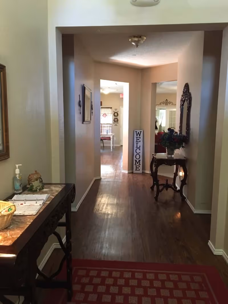 A hallway in Silver Cottage Senior Living with wooden flooring and beige walls. On the left side, there is a small table with a basket, hand sanitizer, and some papers. On the right side, there is a decorative table with a vase of flowers and a large ornate mirror above it. At the end of the hallway, a vertical sign reads 'WELCOME'. The hallway leads to a room with a window and some furniture visible.
