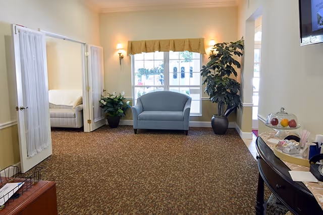 A bright senior living lobby with a blue loveseat beneath a window, potted plants, and a refreshment table to the right.