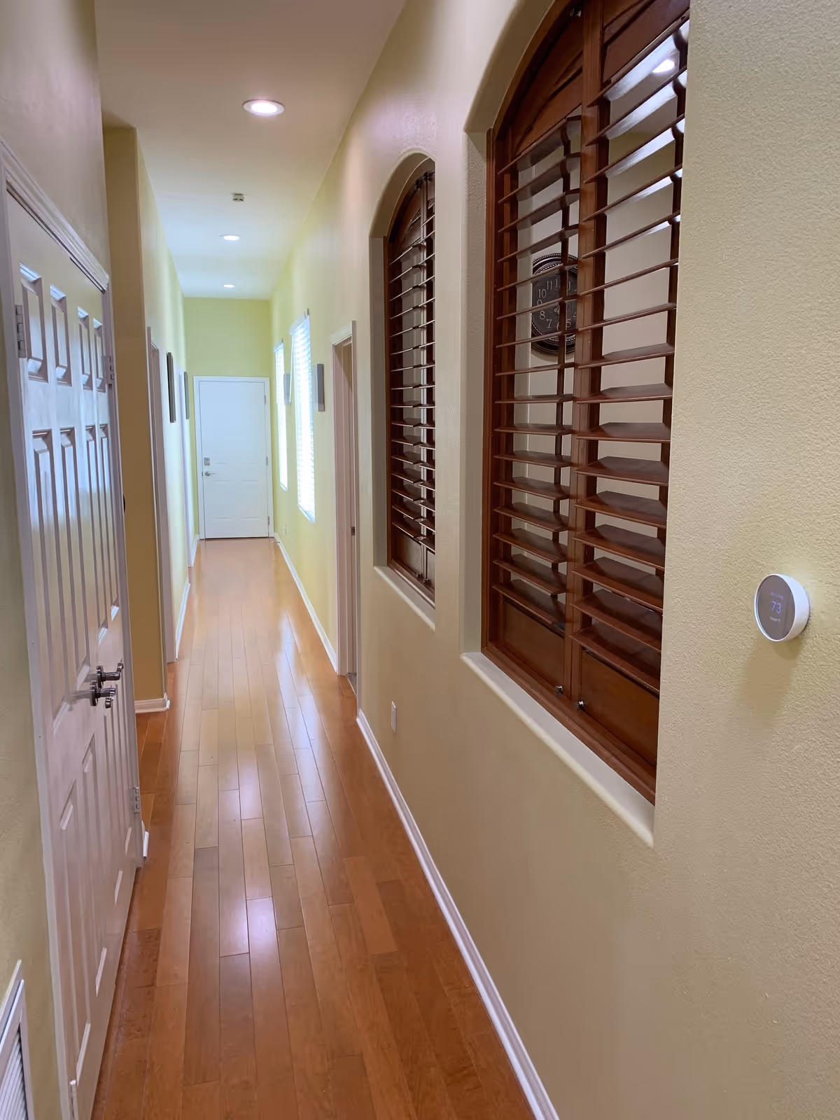 A long, narrow hallway with light yellow walls and wooden flooring. On the right side, there are two windows with wooden shutters and a round thermostat mounted on the wall. On the left side, there are several closed white doors. The hallway is well-lit with recessed ceiling lights and natural light coming from a window near the end of the hallway.