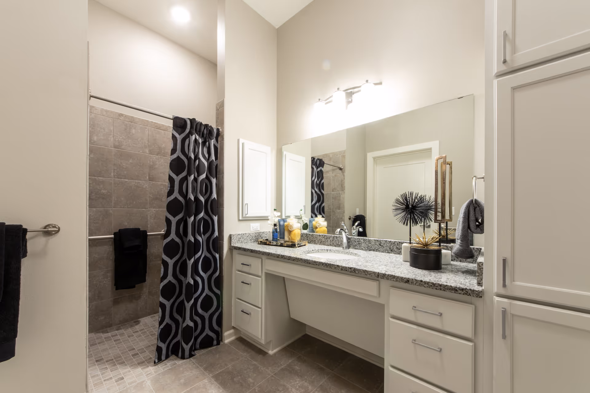 Well-lit bathroom with a granite countertop vanity, large mirror, decorative accents, and a tiled walk-in shower with a patterned curtain.