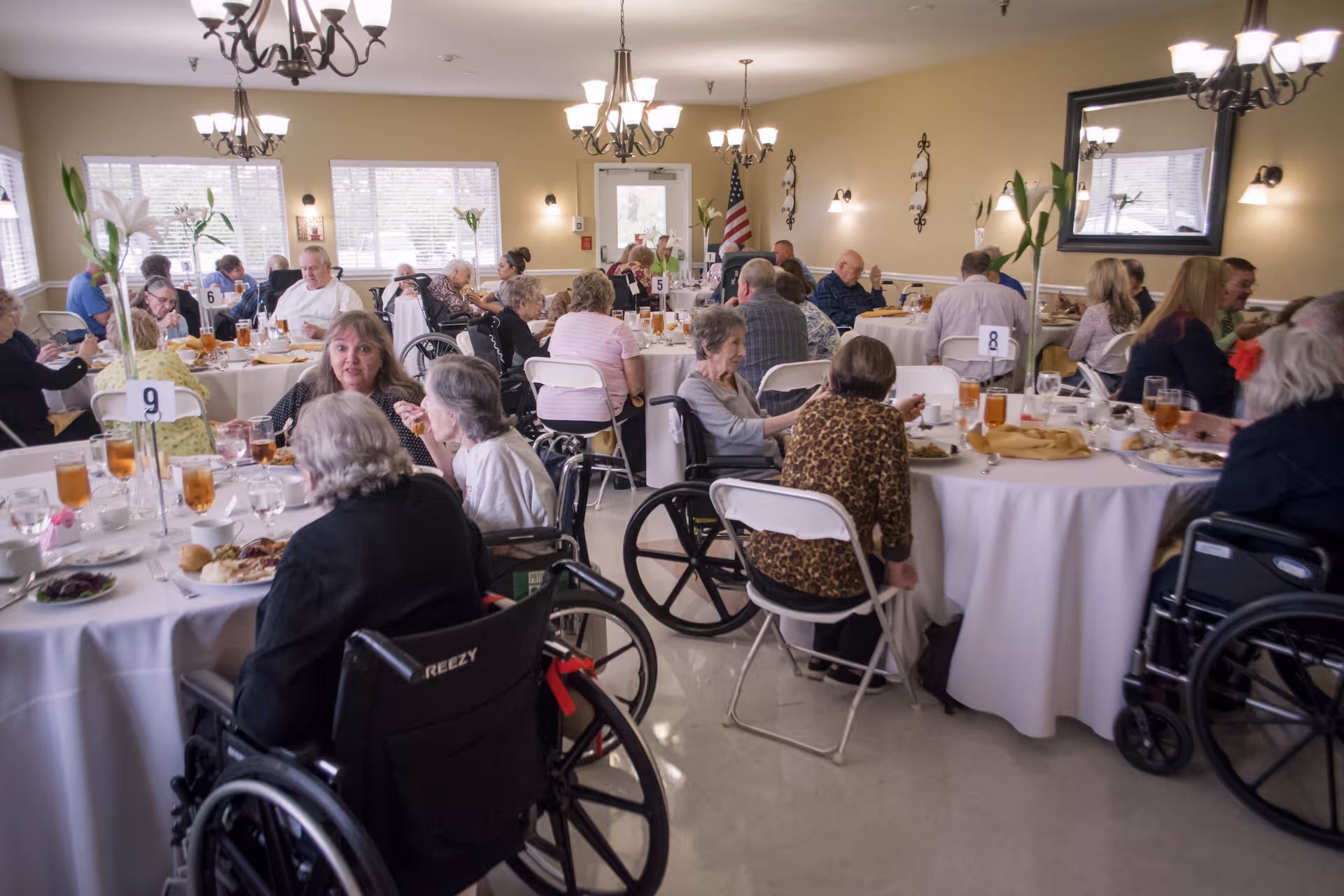 A dining room filled with elderly people sitting at round tables covered with white tablecloths, enjoying a meal together. Some individuals are seated in wheelchairs, and the room is well-lit with chandeliers and wall sconces. There are windows letting in natural light and an American flag near the back door.