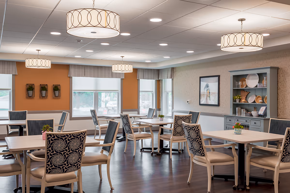 A bright and spacious dining room with multiple square tables and cushioned chairs featuring floral patterned backs. The room has large windows with valances, modern circular ceiling lights, and a gray cabinet displaying plates and decorative items. The walls are painted in warm tones with some framed artwork and small potted plants on the tables.